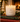 A lit double-wick candle in a frosted white glass jar sits on stacked books, surrounded by pine branches, gold ornaments, and a blurred Christmas tree in the background. The label reads LINNEA and JULETIDE.