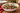 A large white ceramic holiday salad bowl with a red scalloped rim and green berry leaf pattern, filled with cranberry salad, set on a wooden table with plates and pine branches in the background.