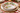 A white ceramic holiday serving bowl with a red rim and green holly leaf pattern, styled on a woven placemat over a charger plate at a festive table setting.