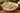 A white ceramic holiday bowl with a red rim and green floral pattern, filled with chocolate chip cookies and topped with cranberries, set on a wooden table with Christmas greenery and lights in the background.