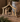 Hand-carved wooden nativity scene figurine with gold leaf accents, displayed on a rustic wooden surface with a blurred decorative pot in the background.