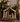A rustic wooden nativity scene figurine displayed on a shelf, featuring Mary, Joseph, baby Jesus in a manger, and a star-topped arch roof. The scene is styled with books, evergreen branches, and a brass lantern in a warm, cozy indoor setting.