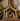 A rustic nativity scene figurine with a weathered wood look and metallic gold-toned trim, displayed on a shelf in front of green garland with warm string lights.