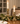 Two lit white taper candles in ornate distressed brass candlesticks arranged on a wooden dining table with greenery and white plates in a softly blurred indoor setting.