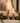 Two brass candlestick holders with lit white taper candles, styled on a mantelpiece with green garland, a framed landscape print, and a gold-rimmed mirror.