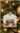 A Christmas ornament shaped like a house hangs on a decorated Christmas tree. The ornament features a rustic nativity scene with a manger, baby Jesus in blue wrapping, and a white sheep, suspended by a gold wire hanger with a gold star topper.