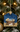 A Christmas ornament shaped like a house hanging from a gold wire loop on a decorated Christmas tree. The ornament features a painted nativity scene with a baby in a wooden manger under a blue starry sky, with a lamb at the bottom.