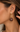 A close-up of a model's ear wearing a gold drop earring with a small pearl-like stud and a dangling oval gold connector leading to a large oval brown stone charm.