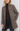 A woman models a brown and beige tweed coat over a dark brown turtleneck and black jeans, photographed against a plain light gray studio background.