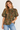 A woman wearing a short-sleeve olive green blouse with a dense multicolor floral print, paired with light blue jeans, against a plain light gray background.