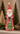 A decorative Santa Claus figurine wearing a red coat and hat with a white beard, holding a green Christmas tree, displayed on a mantel with blurred holiday trees in the background.