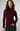 A woman wearing a ribbed mock neck sweater in deep burgundy, paired with black pants, photographed against a neutral light gray studio background.