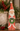 A distressed Santa Claus figurine standing on a tabletop, holding a green wreath. The figurine has a red and white outfit with a weathered finish, set against a holiday background with pine branches, red berries, and stacked books.
