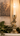 A lit white taper candle in a distressed gold-toned metal candlestick holder, styled on a mantel with green leaves and a framed landscape painting in the background.