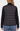 A woman shown from the back wearing a black quilted puffer vest over a black ribbed turtleneck sweater and blue jeans, against a plain light gray studio background.