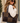 A woman wearing sunglasses and a cream knit sweater carries a large patchwork tote bag with dark brown and taupe leather panels. The brand name 'KATHY LAETON' is visible in gold lettering on the bottom right corner of the bag.