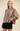 A woman modeling a mocha brown long-sleeve blouse with a gathered high neckline and front buttons, paired with black shorts, against a plain beige studio background.