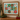 Framed holiday art print featuring 12 decorated donuts arranged in a 3x4 grid on a turquoise background, displayed on a wooden mantel with a gold Christmas tree and books nearby.