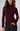 A woman wearing a fitted ribbed mock neck sweater in deep burgundy, paired with black pants, against a light gray studio background.