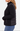 A woman shown in side profile wearing a black ribbed knit and quilted puffer jacket with blue jeans, against a clean white studio background.