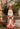 A distressed red and white Santa Claus figurine holding a small red bird, displayed on a wooden surface with holiday greenery, a gold-toned ornament, and books in the background.