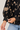 Close-up of a person wearing a black blouse with beige floral embroidery and striped buttoned cuffs, paired with light blue jeans against a light gray background.