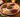 A festive holiday charcuterie board arranged on a scalloped white ceramic platter with a red rim, set on a wooden table with pine branches, a lit candle, and stacked plates in the background.