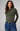 A woman modeling a fitted long-sleeve knit top with a draped asymmetrical neckline in olive green, paired with blue jeans and a brown belt, against a plain light gray studio background.