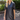 A blonde woman wearing a black short-sleeve faux-leather mini dress with a ruffled V-neckline, standing outdoors on a brick patio with green garden foliage in the background.