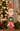 A distressed Santa Claus figurine stands on a wooden surface, holding a green wreath. The background is softly blurred with warm bokeh lights, a Christmas tree, and gold-colored deer.