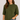 A woman wearing a short-sleeve crewneck knit sweater in olive green, paired with blue jeans, against a plain light gray studio background.