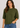 A woman wearing a short-sleeve crewneck knit sweater in olive green, paired with blue jeans, against a plain light gray studio background.