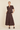 A woman modeling a matching dark brown two-piece outfit consisting of a short-sleeve cropped top with puff sleeves and a high-neckline, paired with a tiered maxi skirt, against a solid light beige background.