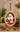An oval Christmas ornament with a gold-toned frame hangs from a curved gold display stand. The ornament features a painted portrait of Santa Claus in his red suit with white trim, set against a warm holiday background with blurred evergreen branches and lights.