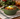 A green ceramic bowl filled with chili, topped with sour cream, shredded cheese, and green peas, set on a plaid napkin with a blurred holiday background and a drink in the background.