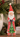 A distressed red Santa Claus figurine holding a green Christmas tree, displayed on a wooden surface with holiday greenery and pinecones in the background.