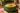 A green ceramic soup bowl filled with creamy cheddar soup, topped with shredded cheese and parsley, set on a wooden table with bread and a plaid napkin.