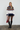 A woman stands against a plain light wall wearing a cream knit sweater with a multicolor Fair Isle pattern across the chest and navy ribbed trim, paired with a black mini skirt, sheer black tights, black knee-high boots, and holding a small red handbag.