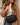 A woman wearing a camel coat and white sweater carries a black crossbody bag with gold hardware and a beige-and-black strap. The bag has a top zip closure and gold logo text reading KATE LEONARDO.