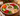 A holiday-themed appetizer platter on a wooden table, featuring a white bowl of creamy dip with red trim, surrounded by fresh vegetables including carrots, cucumbers, snap peas, tomatoes, red bell peppers, broccoli, and tortilla chips.