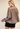 A woman wearing a mocha-brown long-sleeve blouse with a gathered, balloon-style back and black shorts, photographed against a plain beige studio background.