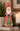 A vintage-style Santa Claus figurine wearing a red coat and hat, holding a green Christmas tree, displayed on a shelf with a lit candle, greenery, and a framed picture in the background.