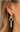 A close-up of a model's ear wearing a multi-layered hoop earring. The earring features a gold huggie hoop, a silver knotted ring, a larger silver hoop, and a dangling silver bar charm.