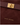 Close-up of a dark brown suede or faux-suede handbag with a matching strap and gold-tone rectangular clasp, featuring the KATIE LOXTON logo in gold near the bottom.