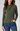 A woman wearing an olive green long-sleeve top with a draped crossover neckline, paired with blue jeans, against a solid light gray background.