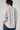A woman shown from the back wearing a white button-up shirt with thin vertical black stripes and gathered ruffle details on the sleeves, paired with blue jeans against a light gray studio background.