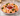A white scalloped serving platter with a red rim holds cooked shrimp arranged around a small central bowl of red sauce, garnished with parsley and lemon wedges, on a marble surface.