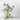 A clear glass bottle with a black pump dispenser containing yellow liquid, styled next to a translucent yellow glass vase with eucalyptus branches on a white surface against a white brick wall.