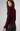 A woman wearing a ribbed long-sleeve turtleneck sweater in deep burgundy, paired with black pants, photographed against a neutral light gray studio background.