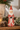 A small Santa Claus figurine in a distressed red and white outfit stands on a wooden surface, surrounded by pinecones, evergreen branches, and a softly blurred indoor background.